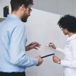 Dos personas que están haciendo un análisis y evaluación del estudio socioeconomico Monterrey, están dentro de una oficina, hay un pizarrón blanco y unos estantes con libros, hay una mujer con cabello chino, lentes y blusa blanca que está de espaldas, esta escribiendo algo en el pizarrón y al lado hay un hombre también de espaldas, con pantalón oscuro, camisa azul claro y lentes que está diciéndole a la mujer que escribir