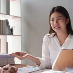 Dos personas sentadas, una mujer con blusa blanca y cabello suelto, enfrente de la otra persona, ella está sentada detrás de un escritorio, sostiene una tabla y en su escritorio hay muchos papeles, porque está realizando recolección de datos de un estudio socioeconomico Monterrey, a su lado izquierdo hay un librero que tiene bastantes libros, pero no está lleno
