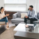 Dos personas dentro de una casa, una mujer y un hombre, la mujer sentada en una silla y el hombre en el sillón, la mujer está haciendo una visita domiciliaria para el estudio socioeconomico Monterrey, la mujer trae pantalón de mezclilla, blusa blanca, tacones y usa lentes, el hombre trae una camisa azul, pantalón azul oscuro y usa lentes, enfrente de ambos hay una mesa de cristal con papeles, hay una alfombra debajo y hay tres ventana detrás de ellos